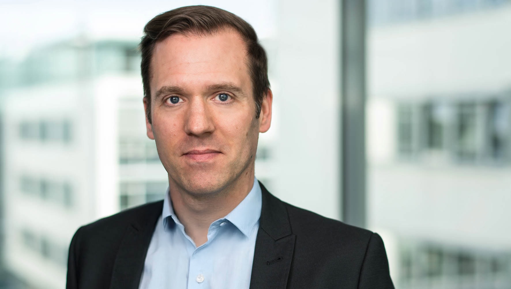 Business portrait of Christopher Kristes in formal attire with a light blue shirt and dark jacket in front of a modern office building in the background. Speaker at the PCI Community Meeting in Amsterdam.