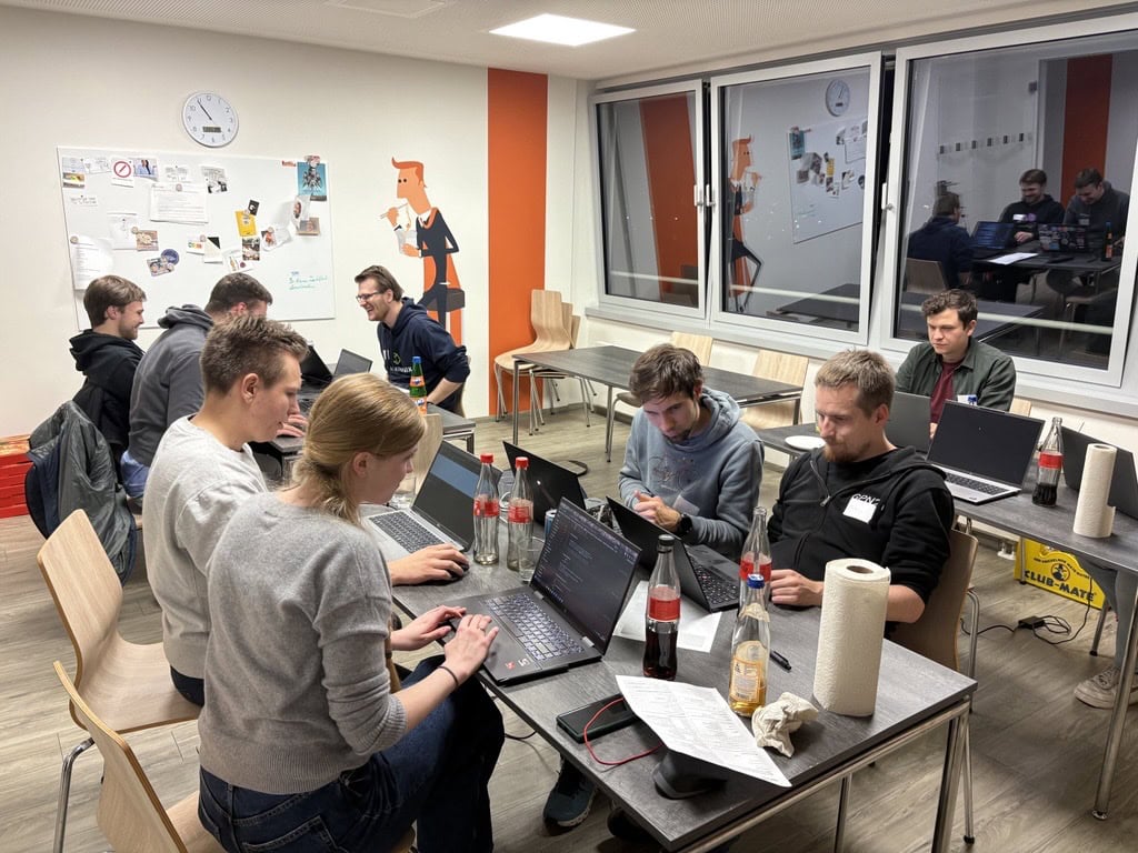 Participants in the usd Hacking Night at the usd office in Cologne work together on laptops, with drinks on the table and a whiteboard in the background.